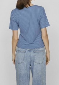 Žena v modrém krátkém ribovém tričku s krátkým rukávem a světle modrých vysokých džínách, ukázaná zezadu na jednobarevném pozadí.