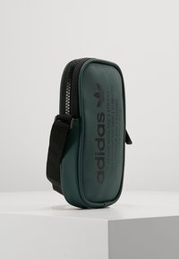 Zelený obdélníkový obal s texturovaným povrchem, vybavený zipem a černým popruhem. Logo "adidas" vytištěné na boku.