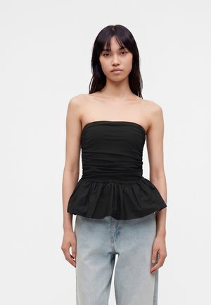 Ung kvinde med langt mørkt hår iført en sort stropløs rynket top med peplum og lyseblå højtaljede jeans, står mod en hvid baggrund.