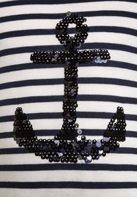 Camiseta de tela a rayas con un gran diseño de ancla de lentejuelas negras en el centro, con rayas horizontales en azul marino y blanco.