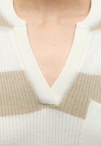 Gros plan d'un pull beige et blanc rayé en maille côtelée avec un col en V profond et une petite poche sur la poitrine.