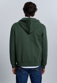 Sudadera con capucha de color verde oscuro, con dobladillo y puños acanalados. Presenta una textura suave y un ajuste relajado. Combinada con vaqueros azules bordados.