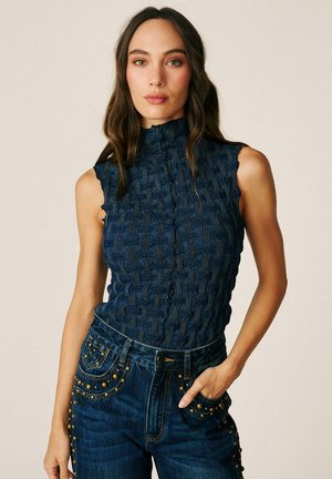 Mujer con cabello largo y oscuro que lleva una camiseta sin mangas texturizada azul marino y vaqueros oscuros con tachuelas doradas, de pie con la mano en el bolsillo.