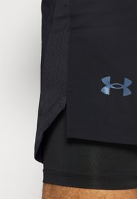 Primer plano de unos pantalones cortos deportivos negros con paneles laterales de malla y el logo de Under Armour, llevados sobre pantalones cortos de compresión negros en la pierna de una persona.