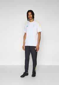 Hvit atletisk t-skjorte med grønne og gule detaljer, korte ermer og brodert logo. Sammen med svarte joggebukser med gule striper.