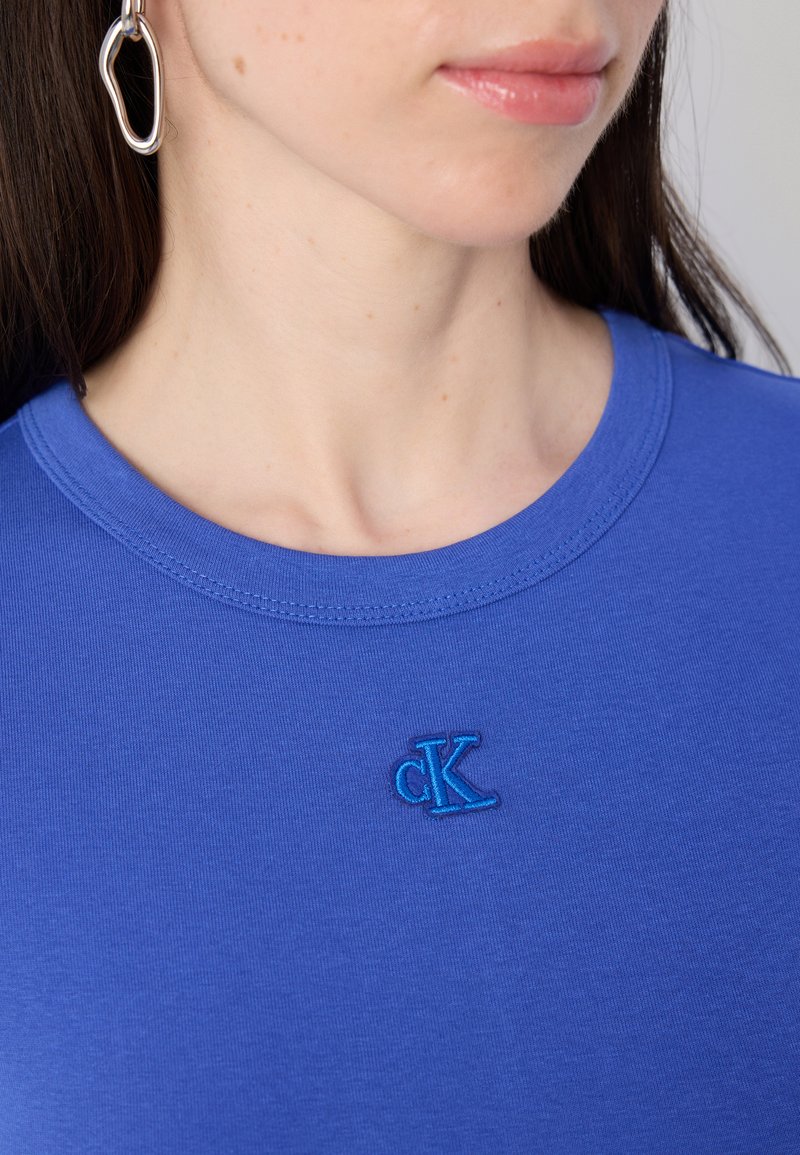 Primer plano del cuello y hombro de una mujer, que lleva una camisa azul con un pequeño logo bordado de "CK" y un gran pendiente de plata.