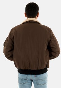 Braune Bomberjacke mit einem beigen Kunstfellkragen, elastischem schwarzen Bund und langen Ärmeln; glatte, glänzende Stofftextur.