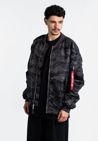 Giubbotto bomber in camouflage grigio scuro e nero con polsini a coste, chiusura frontale con zip e una targhetta con logo sulla manica sinistra. Texture liscia.