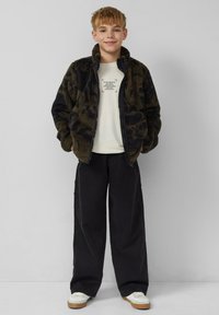 Veste en polaire à motif camouflage sombre, fermeture éclair intégrale, col montant, associée à un t-shirt crème clair et un pantalon noir à jambes larges, baskets blanches.