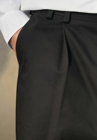 Pantalons noirs ajustés au toucher lisse, avec des plis à l'avant, des passants de ceinture et une poche sécurisée. Associés à une chemise blanche.