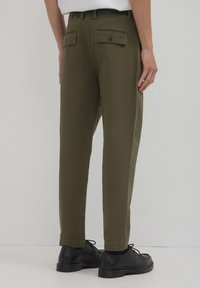 Olivgrüne Hosen mit konischem Schnitt, ausgestattet mit zwei Gesäßtaschen mit Klappen und Knöpfen, aus einem glatten Stoff gefertigt. Getragen mit schwarzen Schuhen.