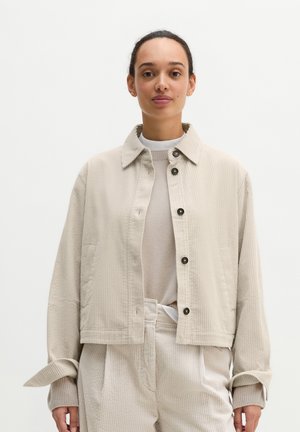 Jonge vrouw met donker haar die een lichtbeige corduroy jas en bijpassende broek draagt tegen een effen lichte achtergrond.