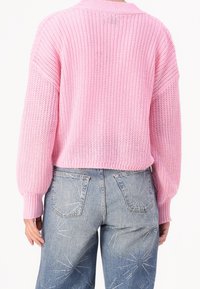 En person klädd i en lös rosa stickad tröja och ljusblå jeans med stjärnexplosion mönster bestående av små nitar på bakfickorna och benen.