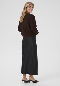 Rjava sweatshirt s rebrastimi manšetami, skupaj z črno maksi krilom z gladko teksturo in subtilnim sijajem, oblečeno z črnimi žabicami na peto.