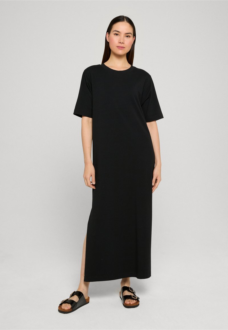 Femme portant une robe maxi noire ample à manches courtes avec une fente latérale et des sandales noires à enfiler, posant devant un fond uni clair.