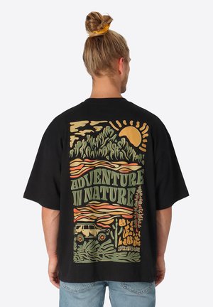 Mann mit blondem Haar zu einem Dutt gebunden trägt ein schwarzes T-Shirt, das auf der Rückseite eine bunte Naturszene mit Sonne, Bergen, Auto und dem Text "Abenteuer in der Natur" zeigt.