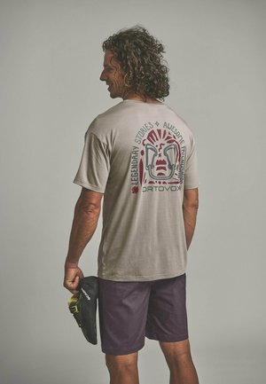 Mann mit lockigem Haar steht mit dem Rücken zur Kamera, trägt ein beiges T-Shirt mit Grafikdruck und dunkle Shorts, hält einen schwarzen und gelben Kletterschuh in der Hand.