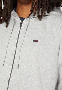 Sudadera con capucha gris con cremallera y textura suave, con un pequeño logo bordado en rojo, blanco y azul en el pecho izquierdo y cordones.