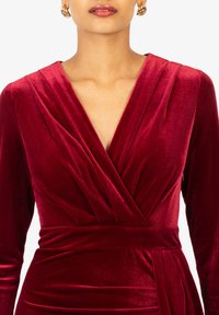 Abito di velluto rosso rubino con profondo scollo a V, maniche lunghe e dettagli drappeggiati in vita, caratterizzato da una texture liscia e morbida.