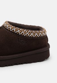 Mocassin en suede marron avec un bord tressé texturé en or et argent le long du col. Dispose d'un logo en relief et d'une semelle en caoutchouc nervurée.