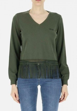 Frau trägt olivgrünes Langarmshirt mit V-Ausschnitt und Fransen am Saum sowie blaue Jeans, steht vor einem schlichten Hintergrund.