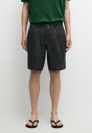 Mann trägt dunkelgraue knielange Shorts mit Knopf und Kordelzug, kombiniert mit schwarzen Flip-Flops und einem grünen Hemd.