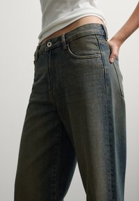 Persona vestida con jeans azul desgastados y una camiseta sin mangas blanca y corta, con una mano apoyada en la cadera.