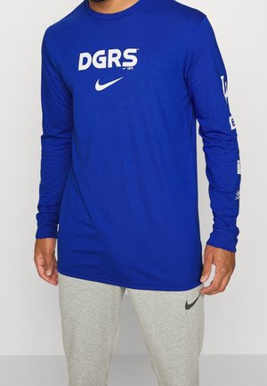 Homme portant un t-shirt bleu à manches longues avec le logo blanc « DGRS » et Nike, associé à un pantalon de survêtement gris clair avec un swoosh Nike noir.