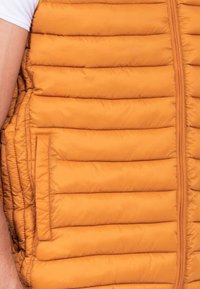 Gilet rembourré orange, avec des sections matelassées verticales et une poche latérale. Conçu en tissu léger et lisse avec une fermeture éclair.