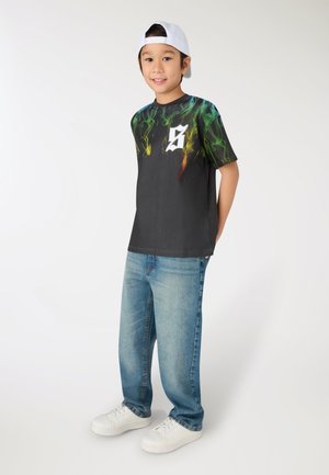 Ragazzo con una maglietta nera con motivi di fumo verde e giallo, jeans azzurri, sneakers bianche e un berretto bianco indossato all'indietro, in piedi e sorridente.