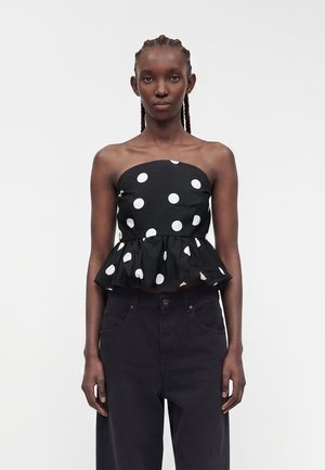 Donna con capelli intrecciati che indossa un top peplum nero senza spalline con pois bianchi e pantaloni neri a vita alta su sfondo semplice.