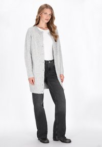 Grauer gestrickter Cardigan mit Knopfverschluss vorne, langen Ärmeln und geripptem Saum, getragen über einem weißen Hemd und kombiniert mit schwarzen weiten Jeans.