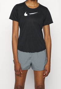 Persona con una camiseta negra de Nike con un logo de swoosh en capas y pantalones cortos deportivos grises frente a un fondo blanco liso.