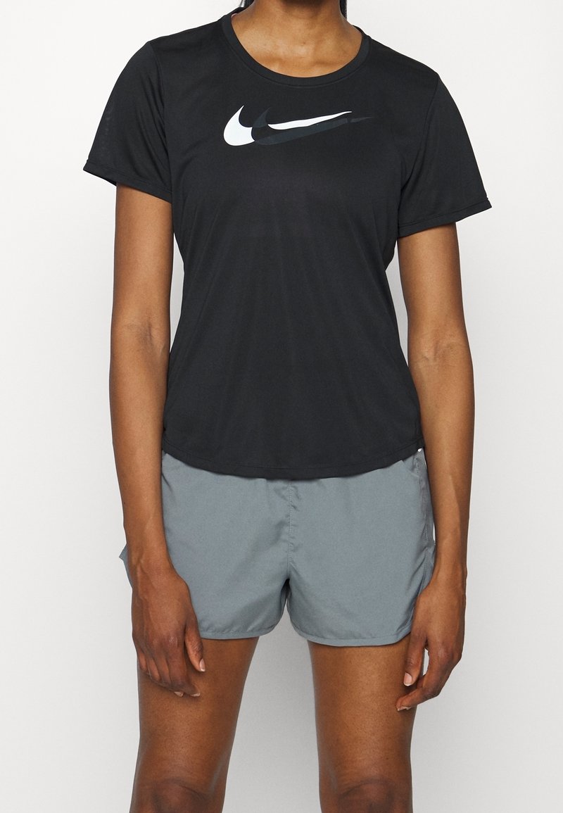 Persona con una camiseta negra de Nike con un logo de swoosh en capas y pantalones cortos deportivos grises frente a un fondo blanco liso.