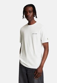 Camiseta blanca de manga corta con cuello redondo, que presenta un pequeño logo negro "LYLE & SCOTT" en el pecho y un logo amarillo en la manga.