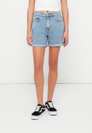 Personne portant un short en jean bleu clair retroussé, un top court blanc et des baskets noires et blanches, debout devant un fond blanc uni.