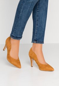 Hnědé semišové lodičky na vysokém podpatku se špičatou špičkou, vlnitými okraji a štíhlým, průhledným podpatkem, kombinované s modrými zahnutejšími džínami.