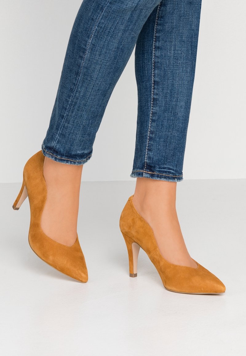 Hnědé semišové lodičky na vysokém podpatku se špičatou špičkou, vlnitými okraji a štíhlým, průhledným podpatkem, kombinované s modrými zahnutejšími džínami.