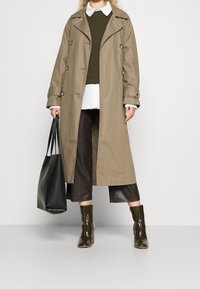 Gabardina beige con botones de doble botonadura, superpuesta a un suéter verde oliva y una camisa blanca, combinada con pantalones cortos marrones y botas de tobillo brillantes.