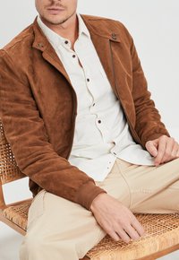Giacca bomber in suede marrone con chiusura a zip, bottoni a pressione, polsini a coste e una texture liscia, indossata sopra una camicia chiara.