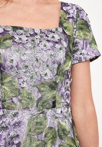 Lila blommig klänning med gröna broderade blad och blommor, fyrkantig halsringning och korta ärmar med texturerade tygg detaljer.