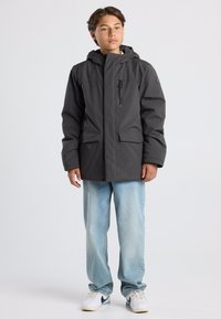 Harmaa eristyksellä varustettu takki, jossa on huppu, sisältää vetoketjullisen rintataskun ja kaksi etutaskua; päällä vaaleansiniset farkut ja valkoiset lenkkarit.