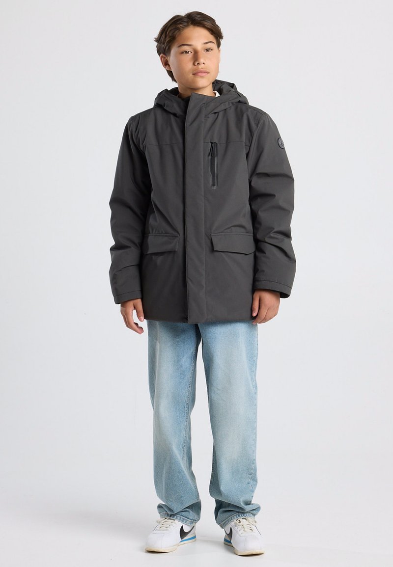 Harmaa eristyksellä varustettu takki, jossa on huppu, sisältää vetoketjullisen rintataskun ja kaksi etutaskua; päällä vaaleansiniset farkut ja valkoiset lenkkarit.