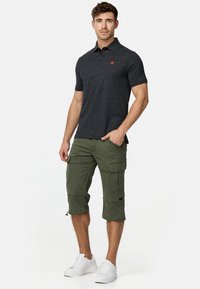 Polo noir avec une texture subtile et un logo orange, associé à un short cargo olive vert avec de nombreuses poches et des ourlets retroussés. Baskets blanches.