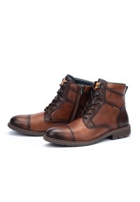 Botas de tobillo de cuero marrón con cordones y cremalleras laterales, con punteras cosidas, mostradas sobre un fondo blanco.