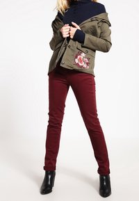 Oliwkowa kurtka z kwiatowym haftem na kieszeni, granatowy sweter dzianinowy, bordo spodnie i czarne botki na kostkę.