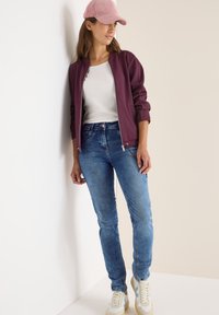 Burgundowa bomberka na białej bluzce, niebieskie wąskie dżinsy i beżowe sneakersy. Ubranie zawiera różową czapkę i jest umiejscowione na neutralnym tle.