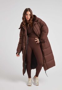 Bruine pufferjas, knielang met een capuchon, met een rits aan de voorkant en zijzakken, gelaagd over een bijpassende bruine strakke outfit.