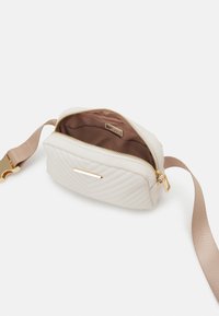 Prošívaná béžová crossbody taška se zlatou kovovou výbavou, zapínáním na zip a hnědým textilním vnitřkem. Nastavitelný popruh a horizontální design.