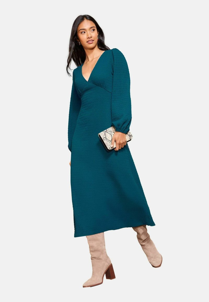 Tamsiai žalia ilgomis rankovėmis su giliu V formos kaklu, tekstūruotas audinys, šoniniai plyšiai, derinama su smėlio spalvos aukštakulniais batais ir raštuota rankine.